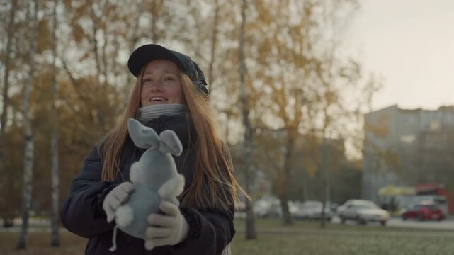 White woman holding plush bunny outdoors, smiling and pretending to snatch toy like a street performer, wearing cap, headphones, scarf and gloves, backpack on shoulders, golden birch trees and parked