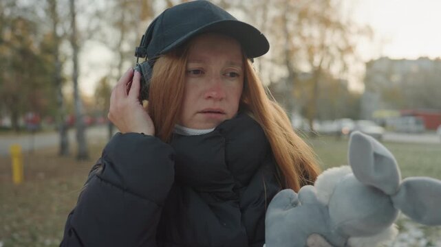 Redhaired woman outdoors holding stuffed elephant, wearing cap and headphones in cold autumn park, scanning surroundings with wary glance, clutching toy while sensing snatcher or prowler nearby,