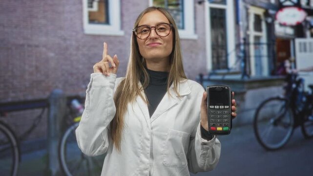 Young woman smiling, holding payment terminal and pointing index finger on street; confidence trust commerce.