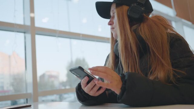 Ginger woman window glance checking phone, student role scanning surroundings while seated by glass, wearing cap and headphones with overcoat, brief turn of head toward lobby, reflective city