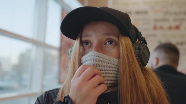 Young white woman with cap adjusting knit mask, headphones on, gazing out city window, tense vigilance against snatcher. Student commuter turns lookout, fingers on scarf, emotional closeup shows