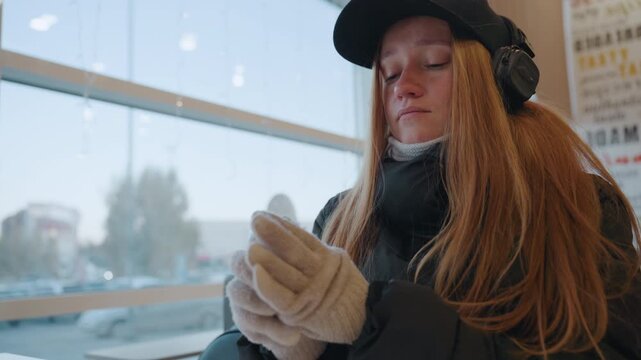 Redhead woman in cafe putting gloves, headphones and cap by window city street outside blurred focused expression as hands slide into wool gloves, breath fogging in winter light furtive scout vibe,