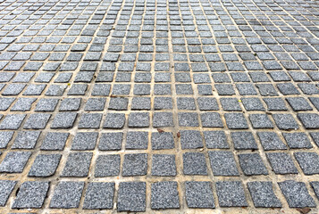 Fototapeta premium Gray stone square tiles arranged in a grid pattern fill the ground surface with sand and small debris between the stones creating a textured and geometric appearance