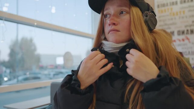 White redhead woman alert in cafe window, headphones on, zipping jacket and adjusting scarf while scanning surroundings for snatcher or pickpocket, cold light through glass, subtle tension