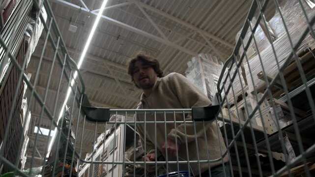 Man places blue paint can into metal cart inside building supply store. Camera looks up from cart basket toward shopper and bucket.