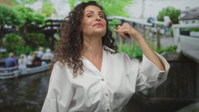 Woman makes phone talk gesture with thumb and little finger on street wearing white shirt near canal boats; communication connection conversation.