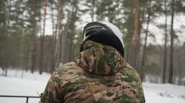 White helmeted rider scans snowy trail ahead, camo jacket and goggles visible as rider surveys pine forest, subtle head turn and throttle check suggest cautious scouting and route planning