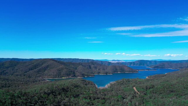 Drone flying alongside Lake Eildon in Victoria, Australia. Side view of blue water with green hills and mountains in daylight, showing calm natural landscape.