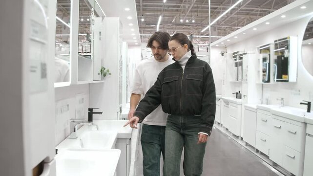 Young family examines white bathroom sink display in hardware store. Couple leans close to check basin and faucet details.
