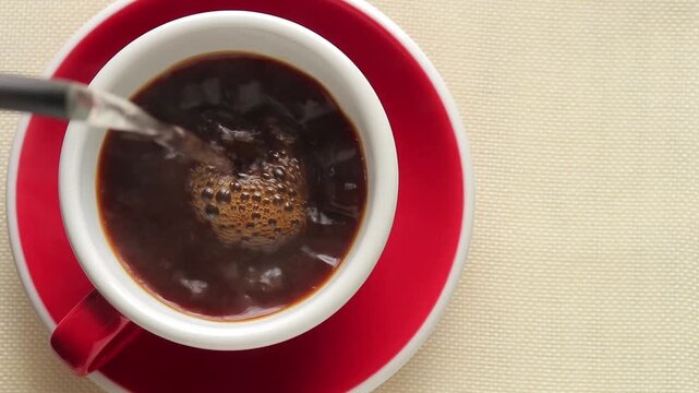 Pouring hot water from a kettle into a red cup with instant coffee powder on a tablecloth.