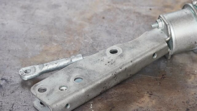 Panning shot of a new metallic turbocharger wastegate actuator and mounting bracket assembly on a weathered workshop surface during an engine rebuild.