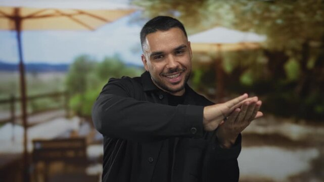 Smiling young hispanic man makes spending money gesture with hands in building with playful smile; generosity abundance wealth prosperity.