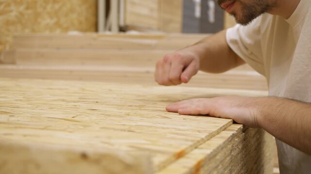 Man examines oriented strand board sheet in hardware store, running hand across surface. He leans down checking texture and flatness of wood panel.
