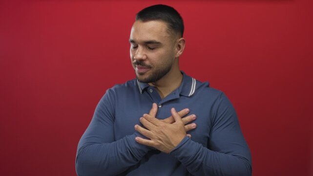 Young hispanic man wearing blue polo shirt smiling and warmly clutching chest in red studio; gratitude.