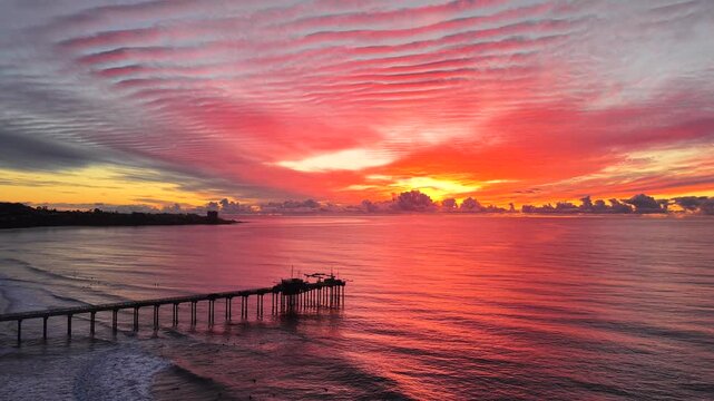 Aerial drone footage of San Diego coast line in Southern California with fly over a pier and stunning sunset sky and red reflections on ocean surfaces.  