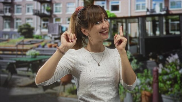 Woman smiling and pointing both fingers forward on an amsterdam street by canal boats and brick row houses, wearing a white eyelet top and visible hands; playful confidence.