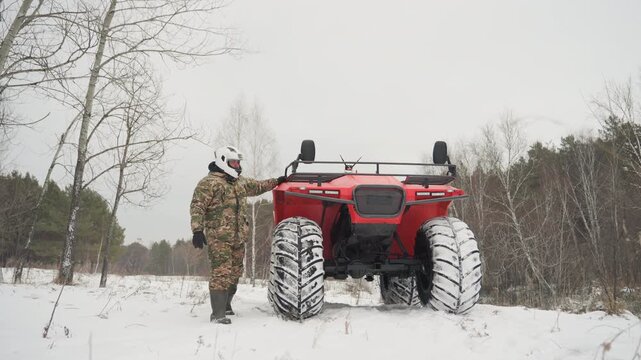 Man in camouflage beside red atv on snowy trail, helmeted rider pauses and inspects vehicle at edge of birch and pine woods visible heavy tires and winter gear convey hunting, offroad maintenance,