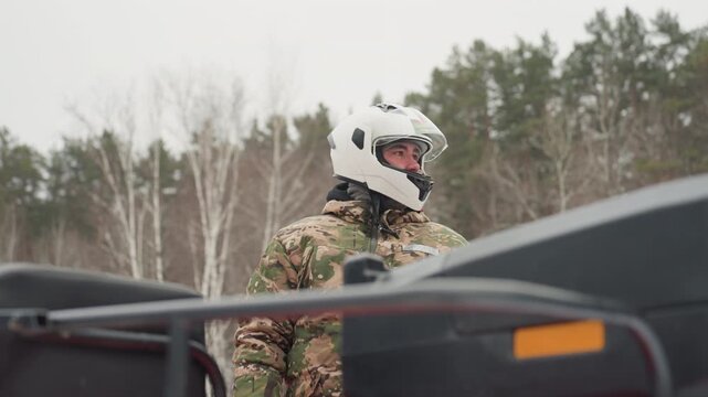 Atv rider in white helmet outdoors standing beside quad in birch forest wearing camouflage jacket under overcast winter sky checking gear and preparing for extreme ride focused expression safety visor