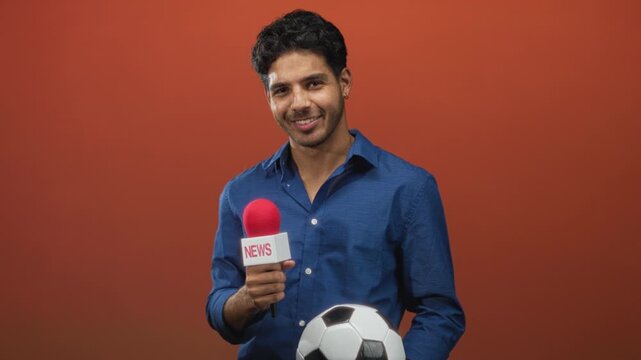 Man smiling holding a red microphone and a soccer ball in a studio shoot; sports interview confidence.