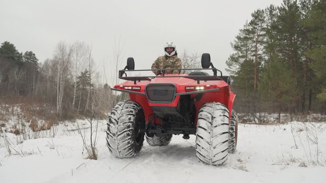 Red atv idling on snowy track, rider adjusting helmet and scanning horizon, chunky tires packed with snow, field edge with sparse trees, muted sky, steady frontal composition, deliberate scouting