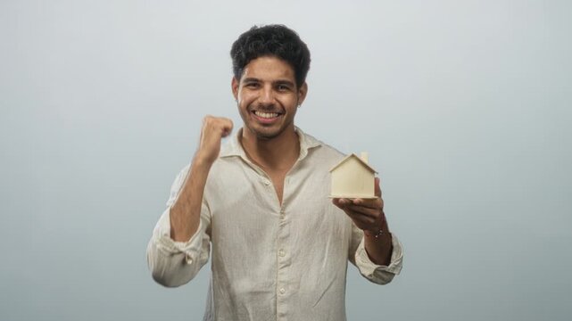 Young hispanic man holding small wooden house model in his left hand while pumping his right fist and smiling toward camera in studio; homeownership joy.