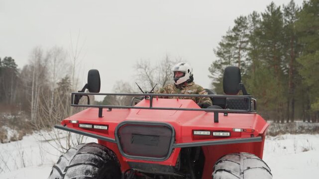 Red atv rider in snowy forest, closeup view of driver wearing white helmet and camouflage jacket steering through powder, aggressive tires kicking snow, engine roar, tense overcast sky and pine