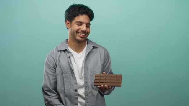 Man holding chocolate bar in hand and smiling while presenting it in studio with teal backdrop and casual shirt; joyful indulgence.