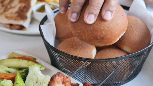 Close up of hand picking up fresh baked bread roll from wire basket on restaurant table during lunch.