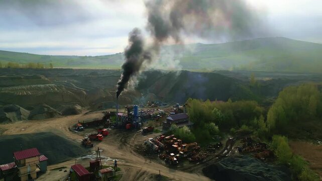 Smoke rises thickly from a mining operation. Vehicles and structures clutter the open pit. Green hills frame the industrial chaos. No people are visible
