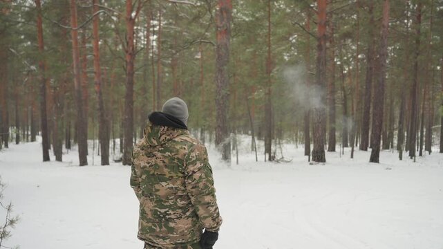 Camouflaged figure walking in snowy forest, breath visible in cold air, tall pine trunks, deep snow underfoot, slow deliberate pace, boot tracks leading into trees, overcast light and muted colors,