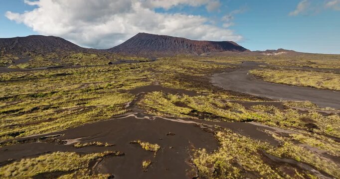 Ambrym Island's vast caldera features the slopes of Benbow and Marum volcanoes rising above a rugged lava field interwoven with green vegetation under a blue sky with clouds. Aerial drone flight
