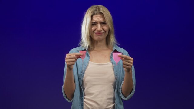 Woman holds two pink menstrual cups comparing sizes with a grimace and pinched fingers in a studio; period choice uncertainty.