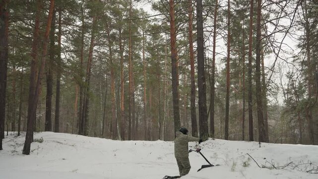 Rider with atv in snowy forest inspecting stalled tracked quad near tall pine trees, light snowfall and muted winter light create quiet, tense recovery scene with visible tracks and bundled gear
