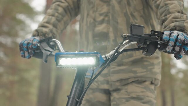 Camouflage rider grips atv handlebar pine forest, bright led bar glares, blue patterned gloves on throttle, camo jacket blends with trees, closeup shows steady control and focused readiness