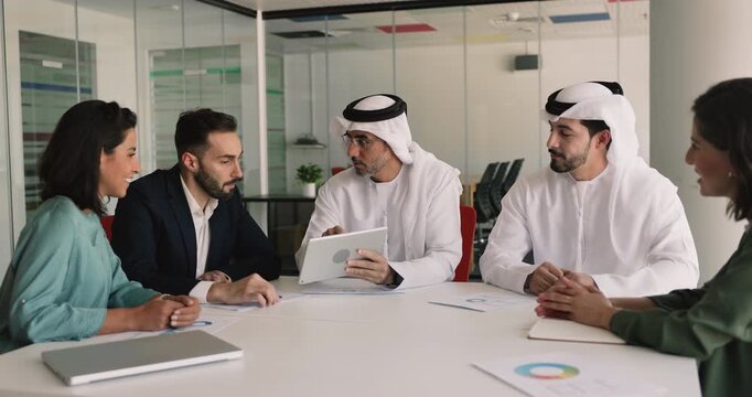 Multicultural team of Arabic and European business partners discussing collaboration terms, reading electronic contract on tablet, shaking hands at meeting table, starting partnership