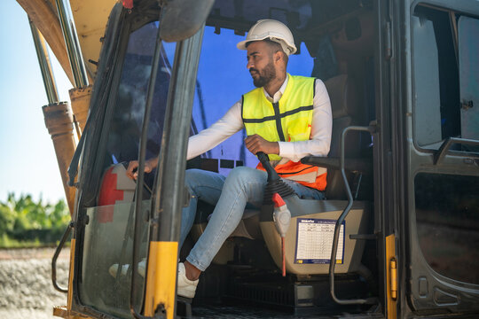 Construction excavator operator man hard hat safety vest cabin control looking through window, seated at lever in heavy equipment, professional driver at construction site under daylight, focus