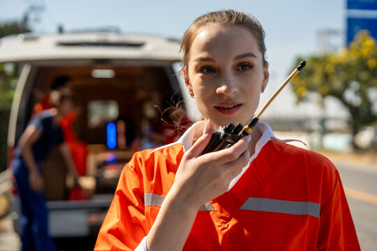 Paramedic emergency ambulance first responder walkie talkie radio uniform orange rescue healthcare on roadside, female young adult in high visibility jacket uses handheld radio near vehicle