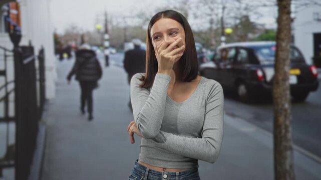 Young hispanic woman covering mouth with hand and slight smile on street, gray top and jeans visible; shy amusement.