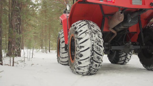 Red ATV tires crunching through snow, closeup shots of aggressive tread packed with snow and mud, front lights glowing, suspension compressing over frozen trail, pine trees framing quiet winter