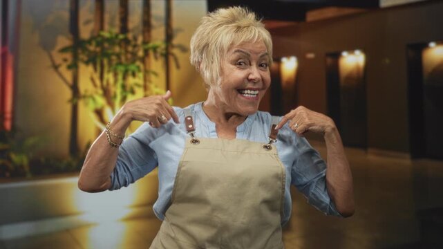 Woman points fingers to apron straps in building lobby, smiling broadly and showing apron; friendly pride.