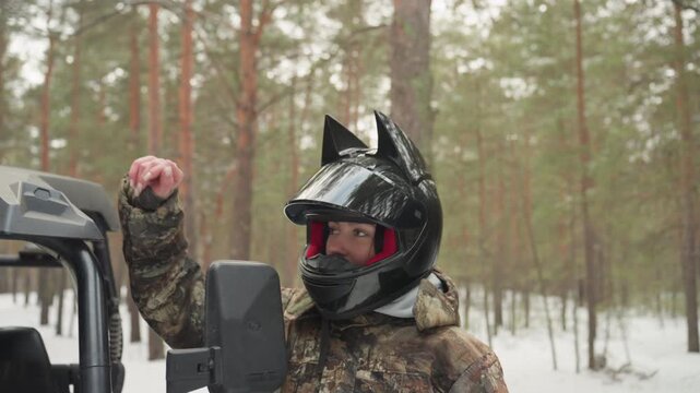 Two riders with helmets beside atv in snowy forest, driver and guide check windshield and engine on camo utility buggy, breath visible in cold air, pine trees and soft falling snow create tense