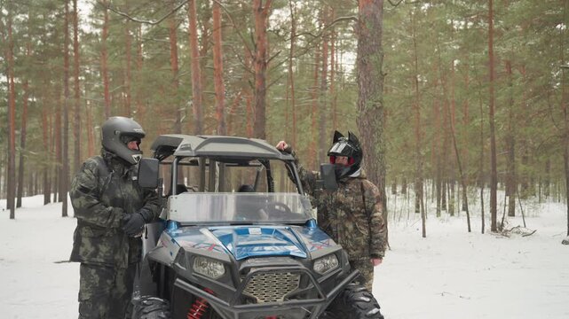 Snowy forest atv extreme ride two helmeted riders beside blue utv in pine woods, camo jackets and goggles, snowcovered ground, closeup of driver face through visor, hands on throttle, prepping