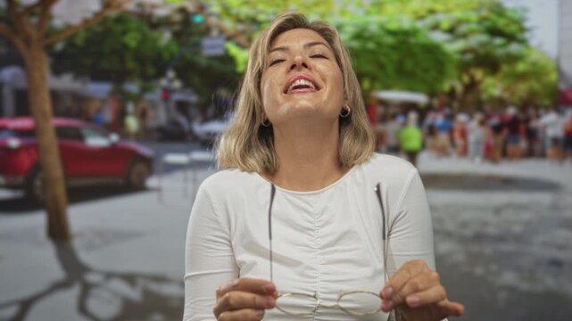 Young blonde woman lifts eyeglasses with both hands on a tree lined city street under sunlight; confidence clarity focus.