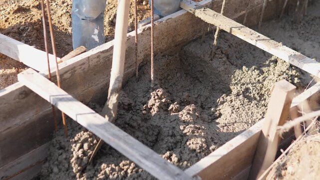 Worker Leveling Foundation Mix With Shovel Inside Wooden Formwork, Boots On Rough Ground, Closeup Of Spade Pressing And Compacting Concrete Near Vertical Rebar, Steady Finishing Motion