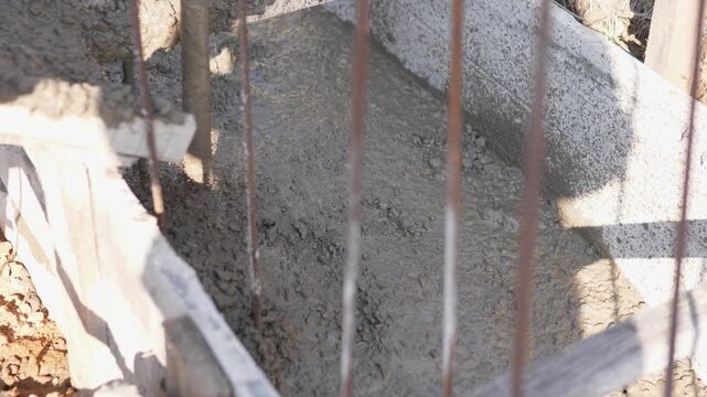 Concrete Being Poured Into Foundation Trench With Wooden Formwork And Vertical Rebar, Sunlight Casting Shadows Across Wet Surface, Site Foreman Inspecting Pour Quality And Alignment