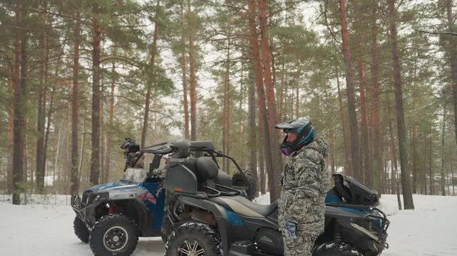 Closeup atv rider adjusts helmet strap and inspects quad controls, camouflage suit and goggles, snowy pine backdrop, focused posture, preparing for extreme offroad ride, safety check and engine start,