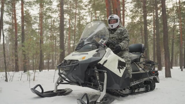 Caucasian rider on snowmobile in forest, winter camouflage jacket and full helmet, closeup lowangle of parked snowmobile on fresh snow, idling engine, tall pine trunks, overcast sky, mood of rugged