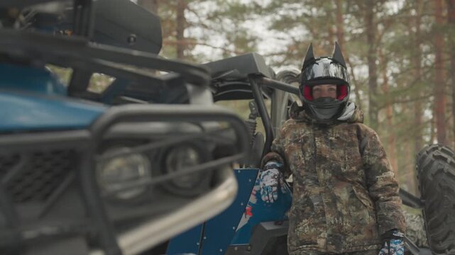 Caucasian rider next to atv front, sharp headlight and blue grill in foreground, blurred helmeted rider in camouflage in pine forest, vehicle inspection and mechanical prep before extreme offroad