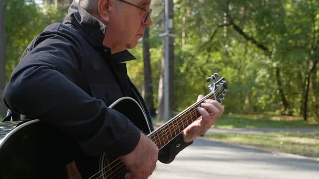 Side profile man playing guitar, white elderly songwriter focused on melody and chords on park path, soft autumn light, casual jacket, thoughtful expression and creative solitude
