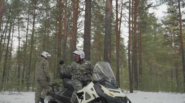 Caucasian men prepping snowmobile in pine forest, camouflaged rider checks engine, companion reviews route, helmets on, cold snow falling, tall pine trunks, fresh tracks in snow, anticipation
